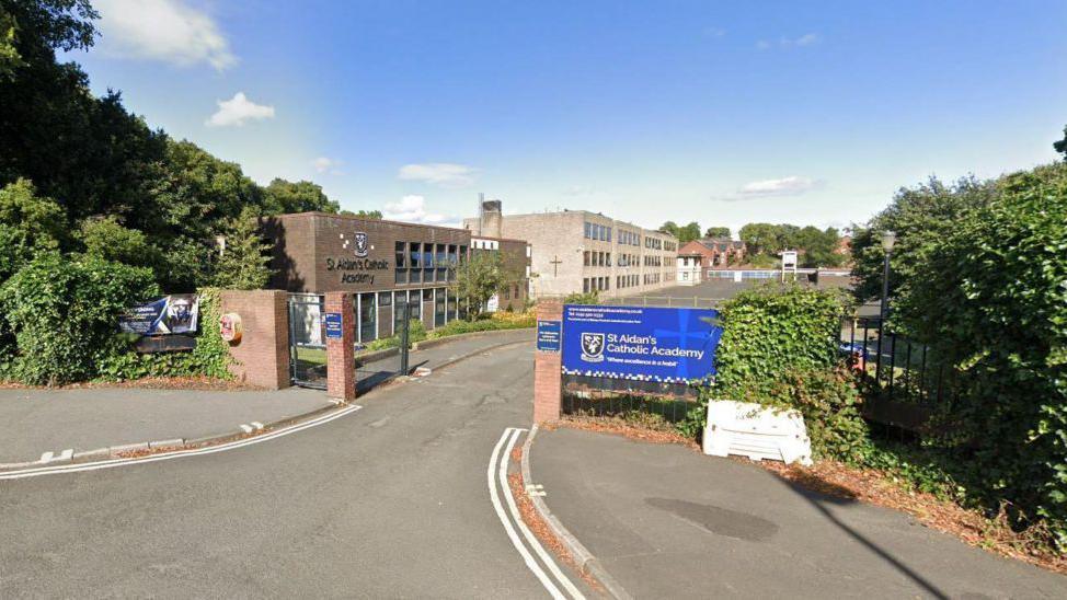 A view of St Aidan's Catholic Academy taken from the road. The school gates are in the foreground. The two school buildings are in the background.