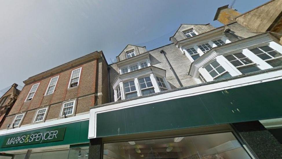 Google Street View looking up at the building facades above the old Marks and Spencer store. The 1930s building on the left is three storeys and the upper floors have sash windows. The Victorian building on the right is four storeys and of pale grey brick. The first floor has two large bay windows, above that are two large sash windows and the top floor has two dormer windows jutting from the roof.