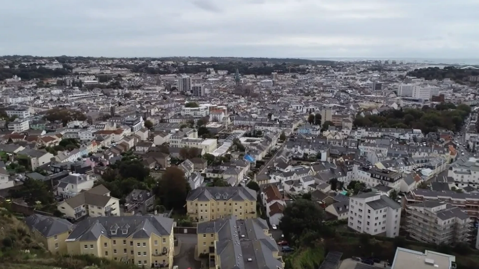 A drone shot of buildings and houses in Jersey. There are green areas with trees and different coloured buildings. There are yellow buildings in the bottom left of the image and white blocks of flats on the right. The sky is grey with clouds.