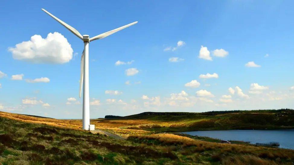 A single white wind turbine in an isolated area of the countryside, with blue skies and some clouds and a lake close to the structure.