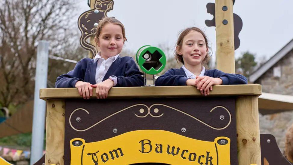 two school girls standing on top of a pirate ship climbing frame.