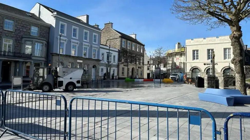 Market square on a clear day, with a road sweeper, and blue barriers in the foreground.