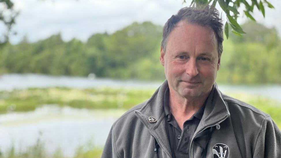 Warden Grahame Hopwood looking into the camera with grey Lincolnshire Wildlife Trust jacket with the reserve in the background