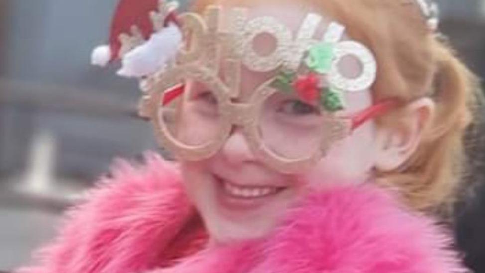 Lila with red hair wearing Christmas glasses which say 'HoHoHo' and a pink fur jacket smiling.