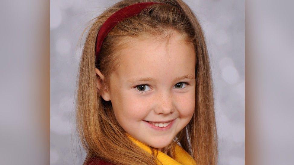 Elsie Dot Stancombe, wearing a red hairband and her school uniform, smiles as she is photographed.