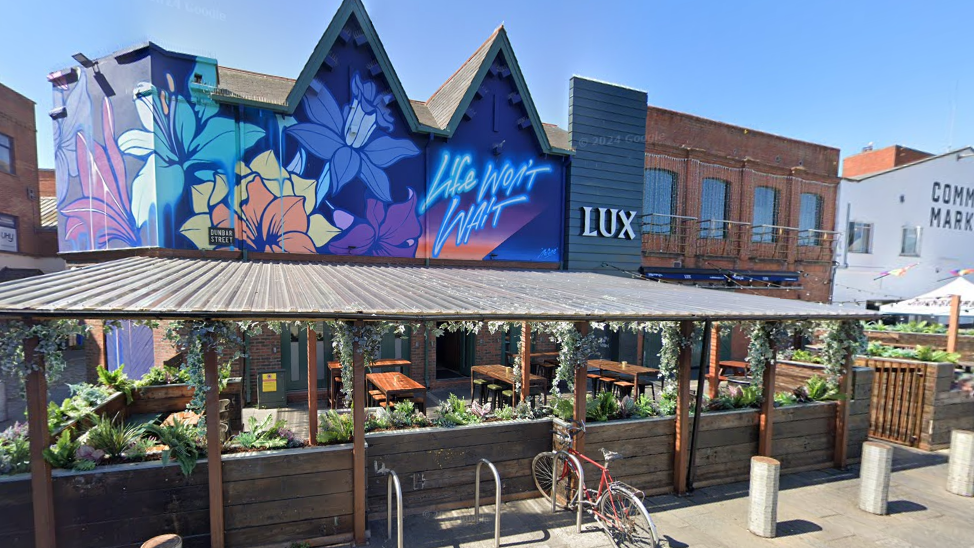 The exterior of Lux nightclub in Belfast. It shows a colourful mural wall with 'life won't wait' in neon-style spray paint. There is also a sheltered beer garden.