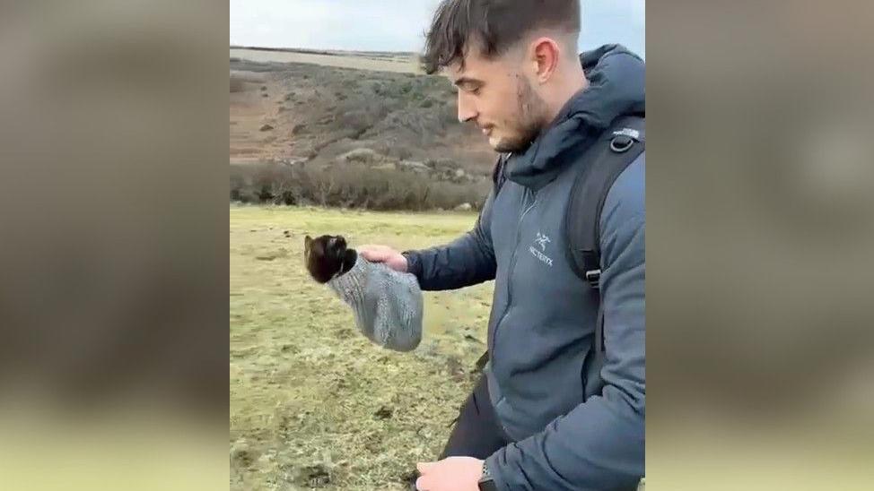 Billy Kelland carrying Bramble in a woollen hat while out on a coastal path with his friends.