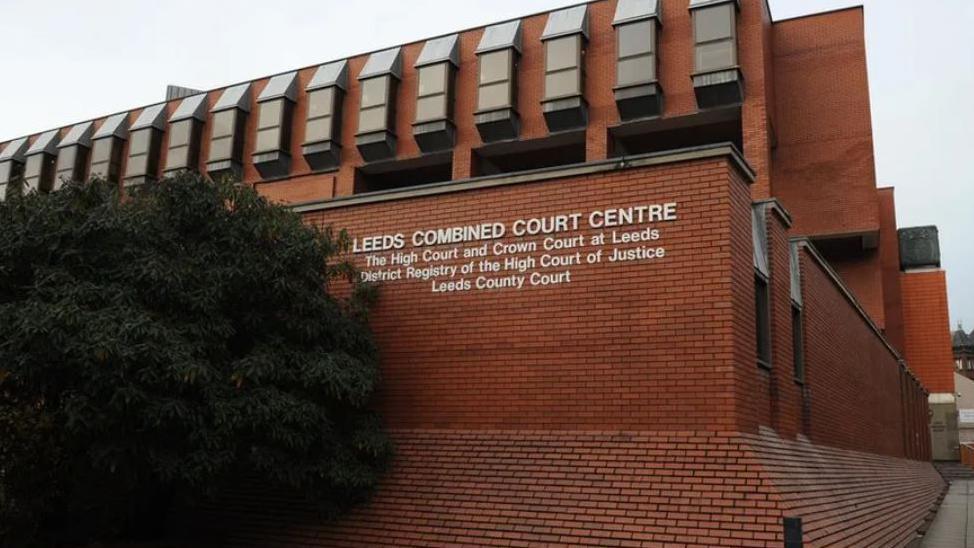 The outside of Leeds Crown Court which is reddish brown. There is a large green bush in the foreground