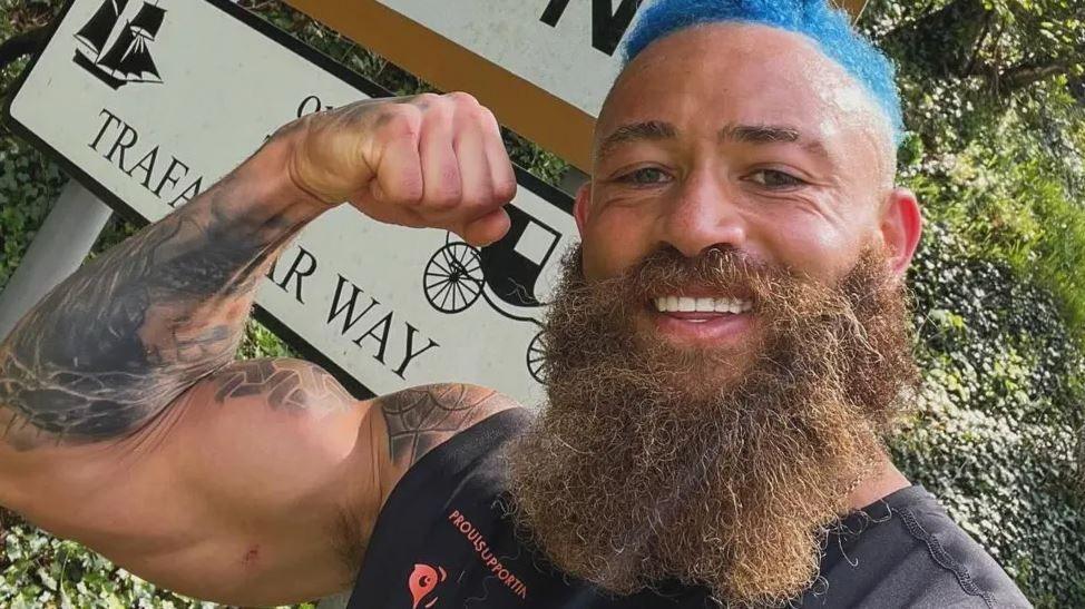 Ashley Cain, flexing his arm while standing in front of a sign that says Trafalgar Way. He's smiling, has a long beard and dyed blue hair