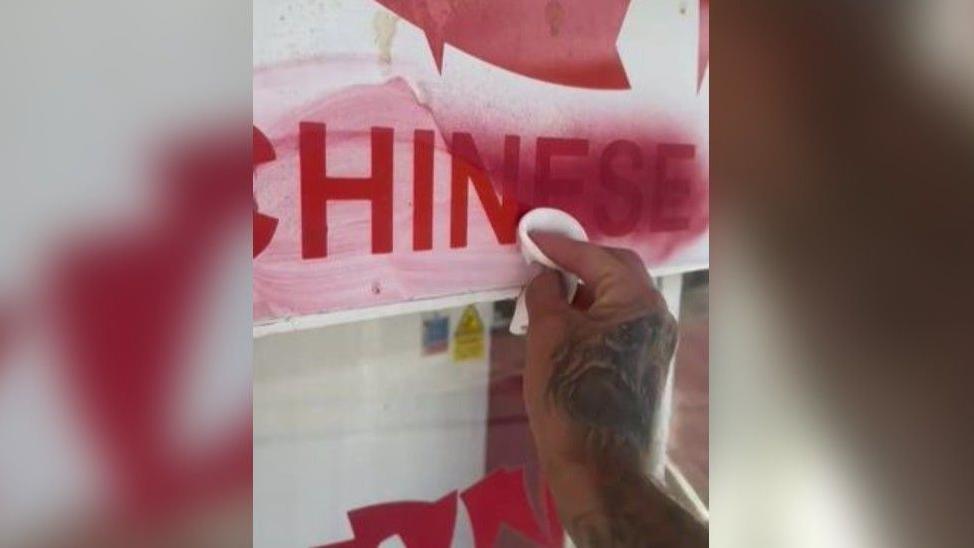 A close up of a man's hand with tattoos cleaning red paint off a sign which says 'Chinese' in red on a white background.