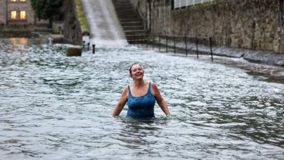 Louise Snelson wearing a blue swimming costume stands in waist-deep water near a slipway. She is looking into the distance and has a big smile on her face. She has a green watch on her left arm.