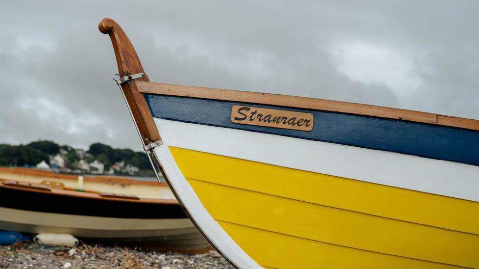 A bright yellow, white and blue rowing boat with the name Stranraer on the side of it
