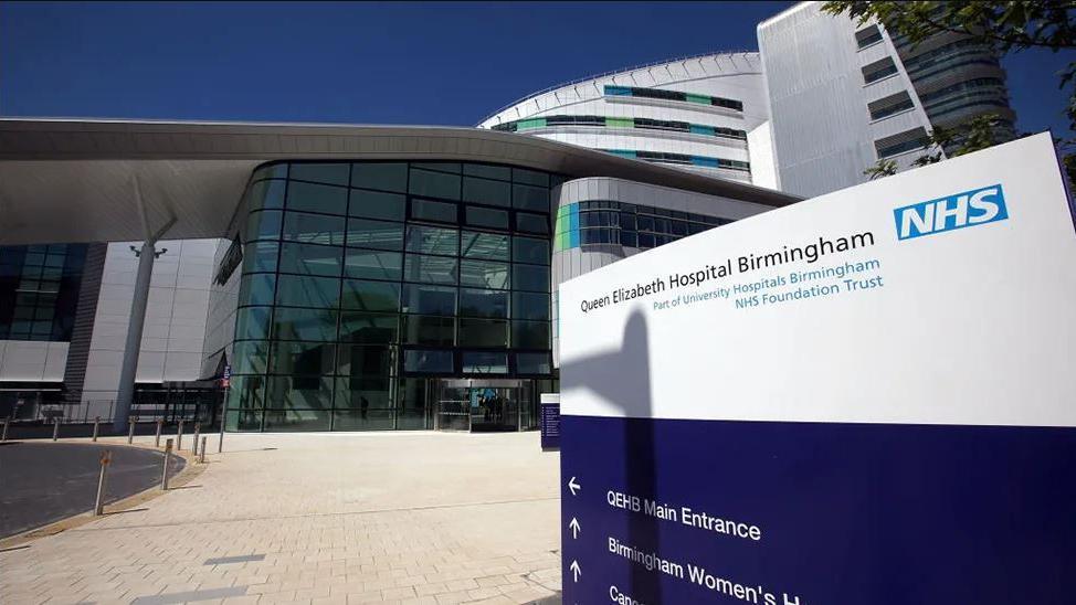 Queen Elizabeth Hospital Birmingham, showing a blue and white sign on the concourse outside, with the main building behind 