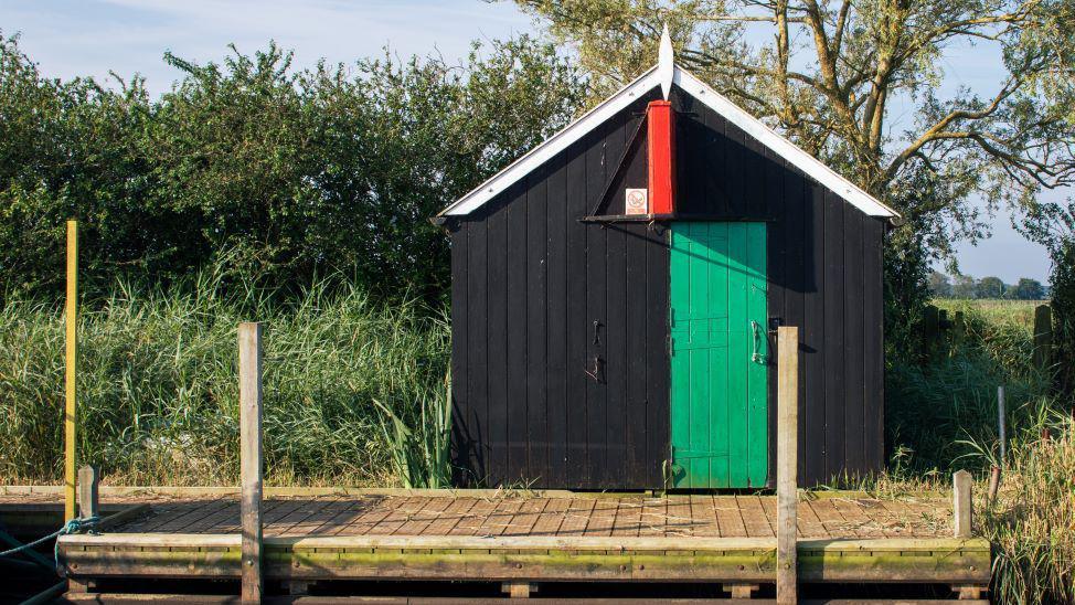 A small black wooden hut. It has a green door and red sign. In front is a small wooden dock. 