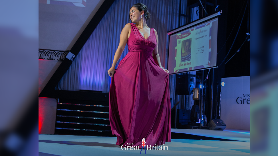 Elle Seline in a long pink dress onstage. She is posing, holding the dress out at each side. There is a projector screen behind her with her name on it and a photo.