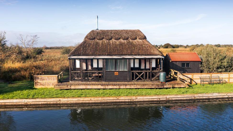 A small black wooden house with a thatched roof on the waterside. The top of the thatched roof is scalloped. It has a wooden deck which wraps around the house. There is a grassy area between the decking and the water's edge.