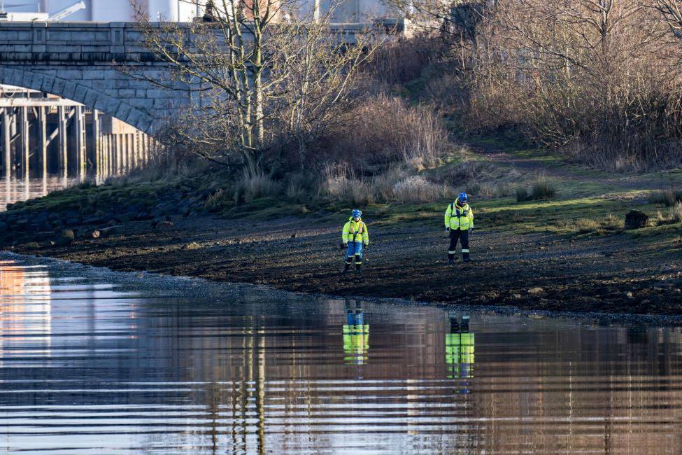 Two searchers in yellow hi-vis jackets on the shoreline of a river, they are reflected in the water, and a bridge arch is in the background.