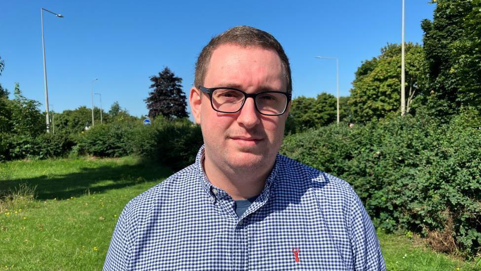 Tom Sisley looks directly at the camera on a sunny day. He has cropped brown hair, wears black rimmed glasses and is wearing a blue and white checked shirt. Behind him is grass and green bushes. 