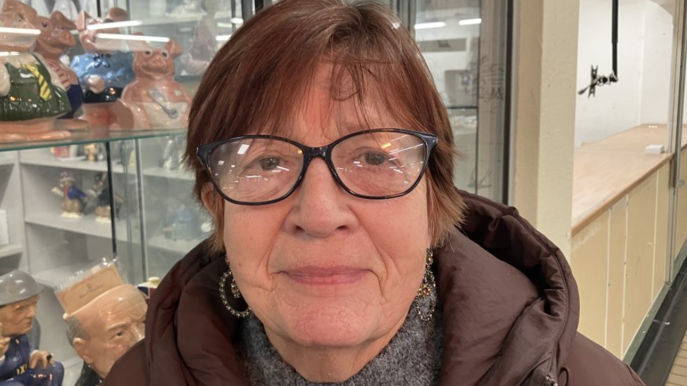 Susan Parniyan standing in front of a market stall. She is wearing black-rimmed glasses and a brown coat. 