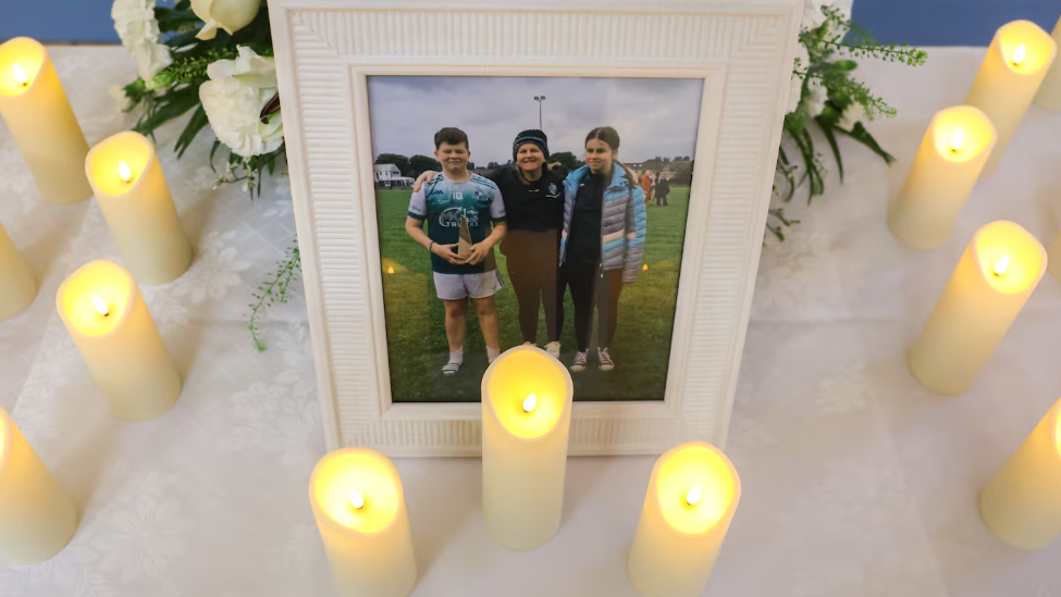 A white table covered in a white linen cloth has a photo of one boy, a girl and a woman. There are lit white candles around the fame. 