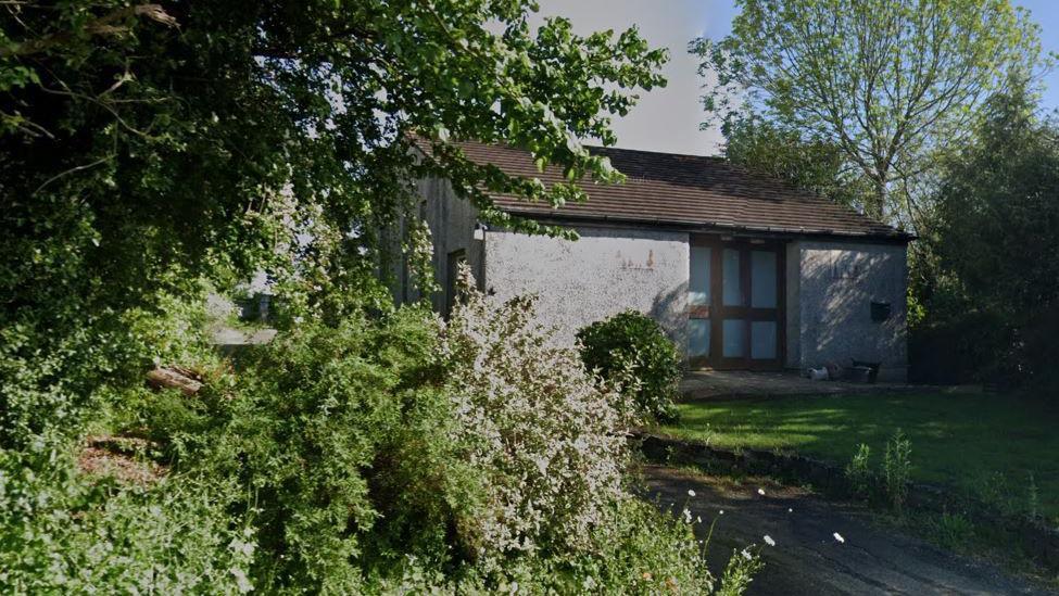 The old Methodist chapel at Crockernwell, on Dartmoor. It is a simple grey, pebble dash building with a wood and glass door in the centre. It is surrounded by trees and shrubbery.