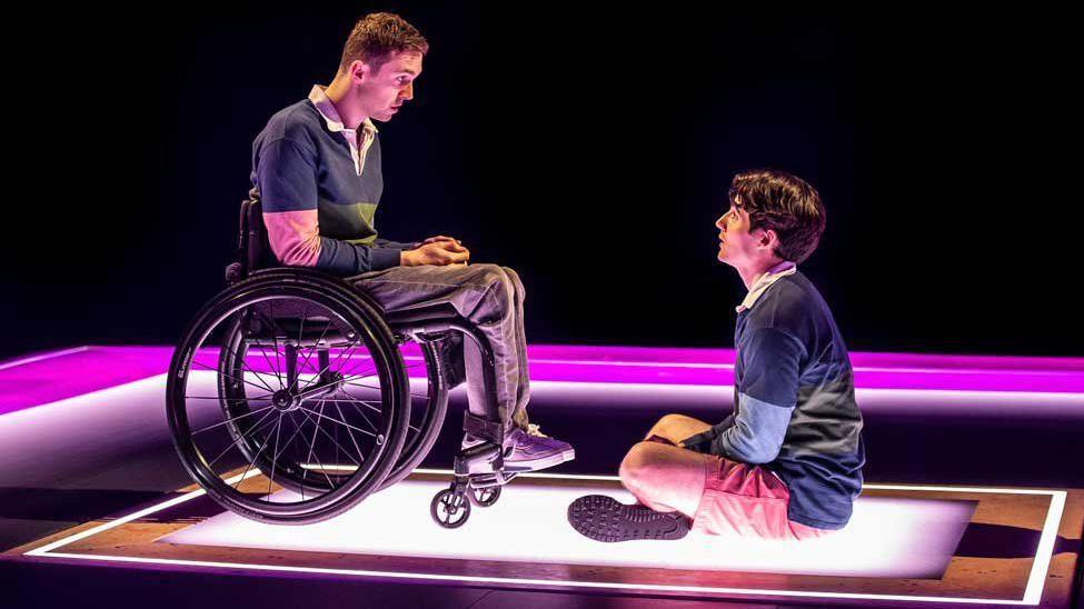 Jonny Amies, wearing a blue rugby shirt and red shorts plays Henry Fraser in his younger years in The Little Big Things. He is sitting cross legged in front of wheelchair user Ed Larkin who portrays Fraser following the accident and is dressed in the same top with grey jeans