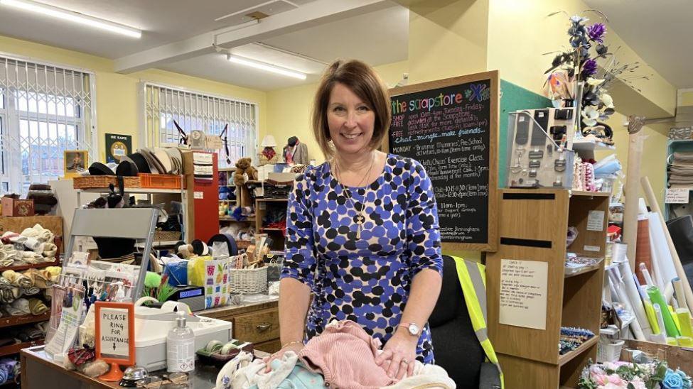 A woman in a blue and black patterned dress in a room with boxes and shelves packed with cloth and other objects