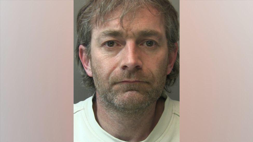 A police head and shoulders shot of a man with grey and brown hair that isn't styled and is nearly at his shoulders. He has a short stubbled moustache and beard and is wearing a white top and looking directly into the camera.