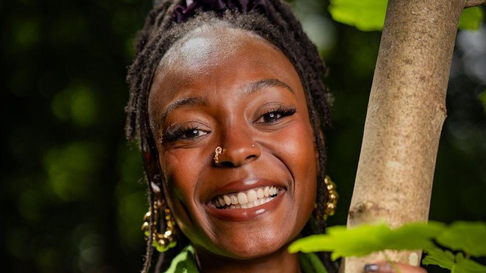 A woman with dark, braided hair smiling into the camera and holding onto a slim tree trunk. She has a gold nose ring and gold hooped earrings.