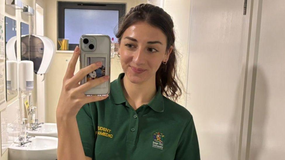 Emilie Griffiths mewn gwisg myfyriwr parafeddygol yn tynnu hunlun