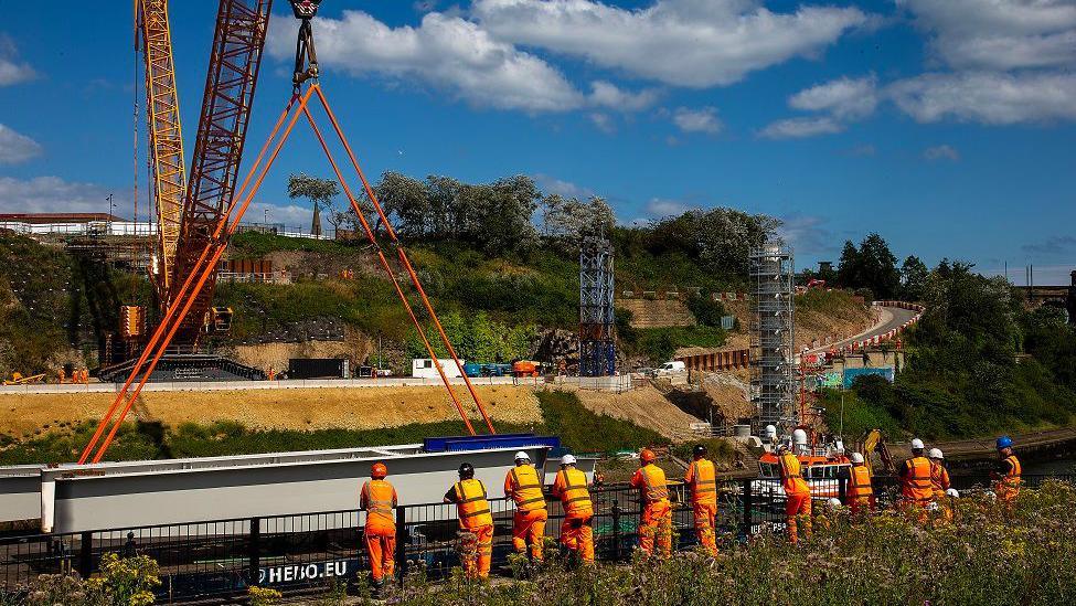 People in orange hi-vis outfits standing on a riverbank look over at a very large crane, carrying a piece of steel. White clouds can be seen in a blue sky overhead. 