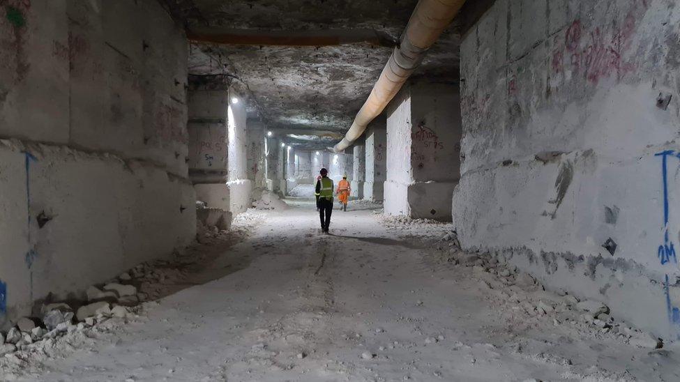 View along a large square horizontal mine shaft. Every surface is white Portland stone. Two men in hi-viz overalls and hardhats are walking away in the distance. The ceiling is about four times their height.