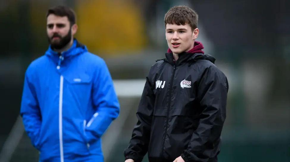 Two male coaches stand in what looks to be a sports pitch, but only one of them - who looks to be younger - is in focus. He has short hair and wears a black coat with the Bristol Barracudas logo. The two men both look ahead attentively as if paying attention to a match unfolding before them.