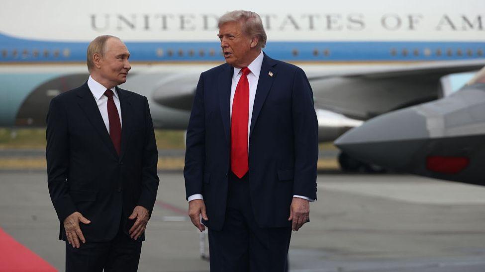 Vladimir Putin, wearing a black suit and maroon tie, standing next to Donald Trump, wearing a blue suit and red tie, in front of Air Force One, with a red carpet seen to the left