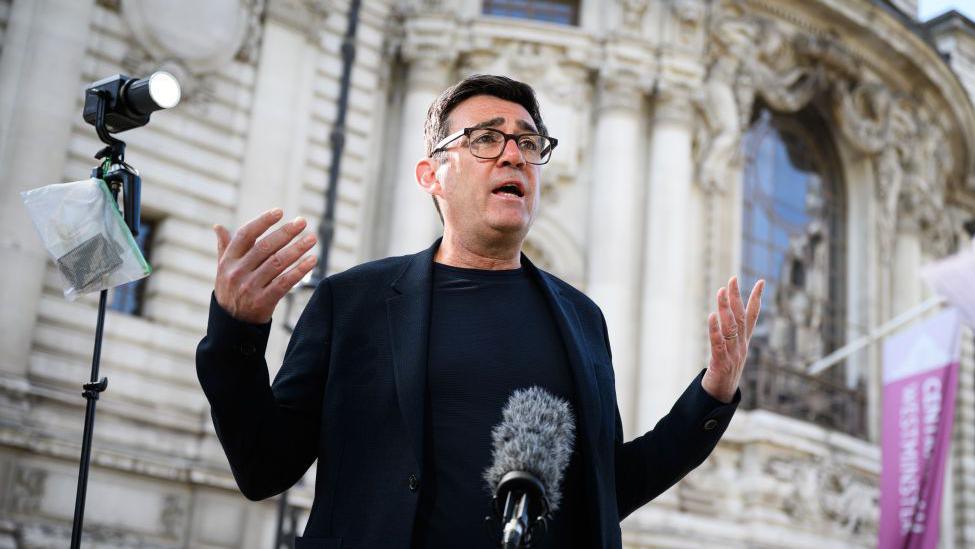 Andy Burnham giving a speech with his arms spread out, with a microphone and a light near him