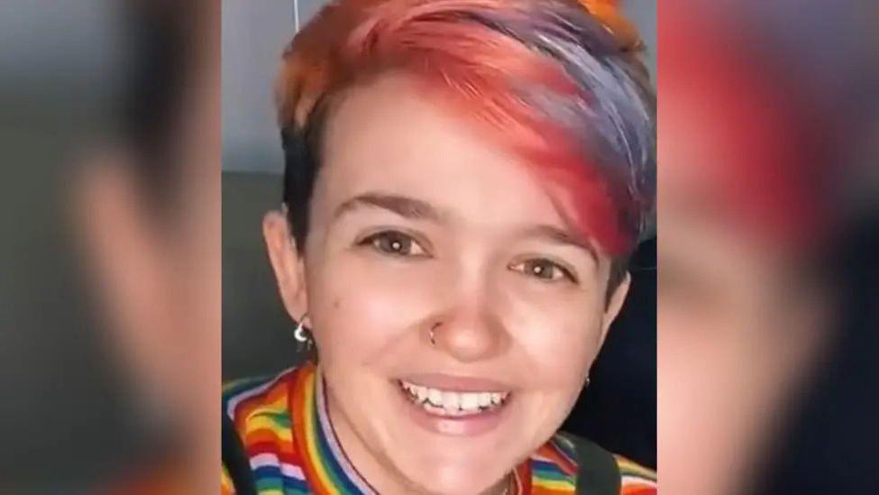 A person with pink and purple hair smiling at the camera. They have a nose ring and are wearing a rainbow shirt.