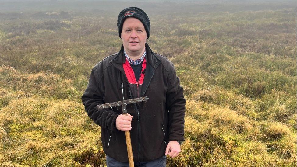 Graham Wilson yn dal cribin mewn ardal niwlog ble mae 'r tir yn drwch o fwsogl