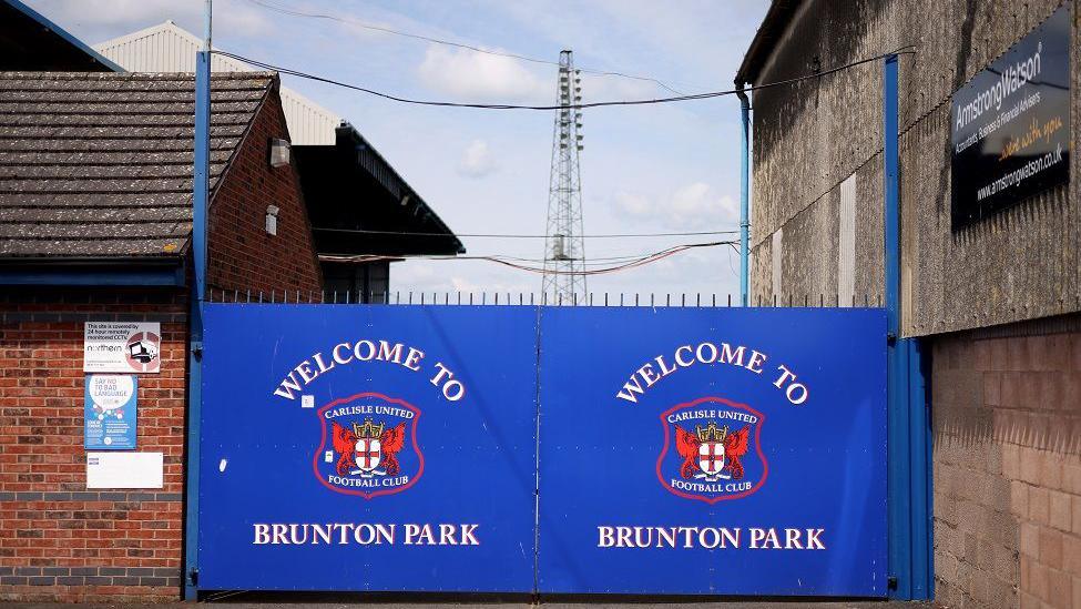 The outside of Brunton Park with a large, blue gate which reads 'Welcome to Brunton Park' alongside Carlisle United's red badge.