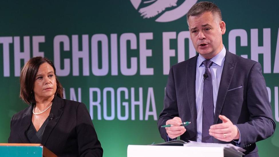 Mary Lou McDonald with Pearse Doherty who is tall with greying dark hair and is wearing a dark suit and speaking at the podium of an election event