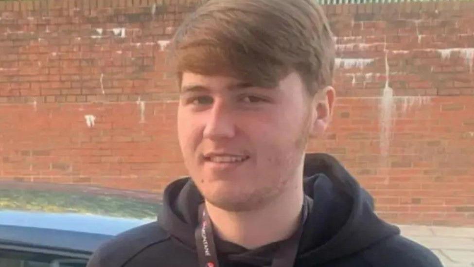 Ellis Cox smiles at the camera wearing a black hoodie in front of a car on a road in front of a red brick wall