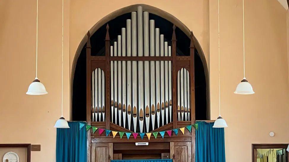 An organ, which blue green red and yellow bunting going across it. There are lights hanging down from the ceiling. To either side of the organ are blue curtains. The wall is a yellow colour.