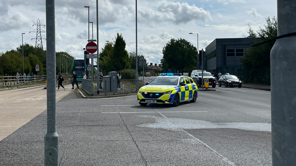 A police car is turning across the road - in the direction of the college which is out of view. It is yellow and black with flashing blue lights on top.