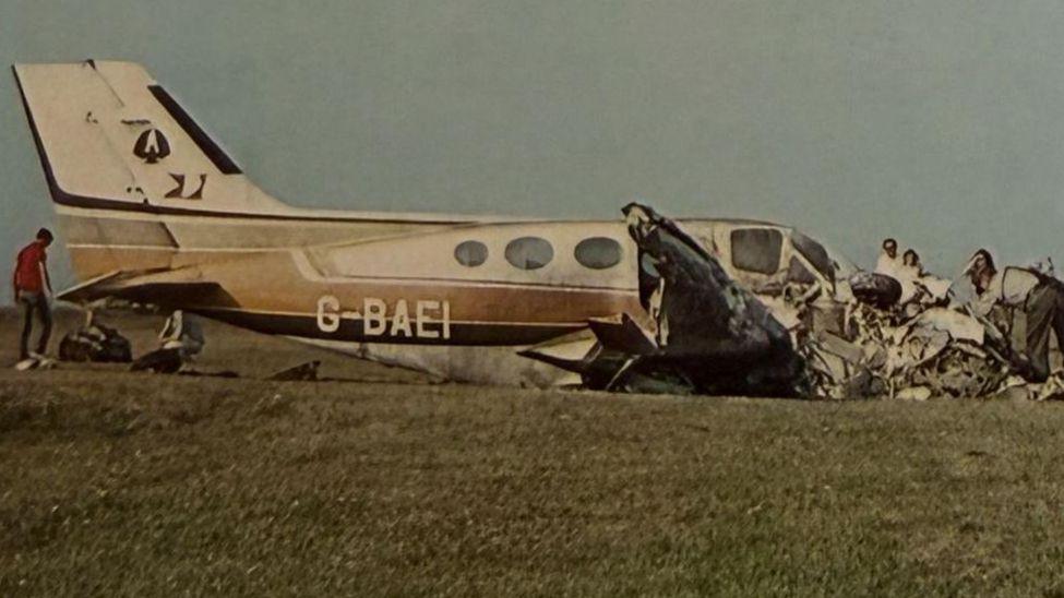 一张旧照片显示，一架小型飞机的残骸停在草地上，人们在挖掘残骸，其他人则在一旁观看。