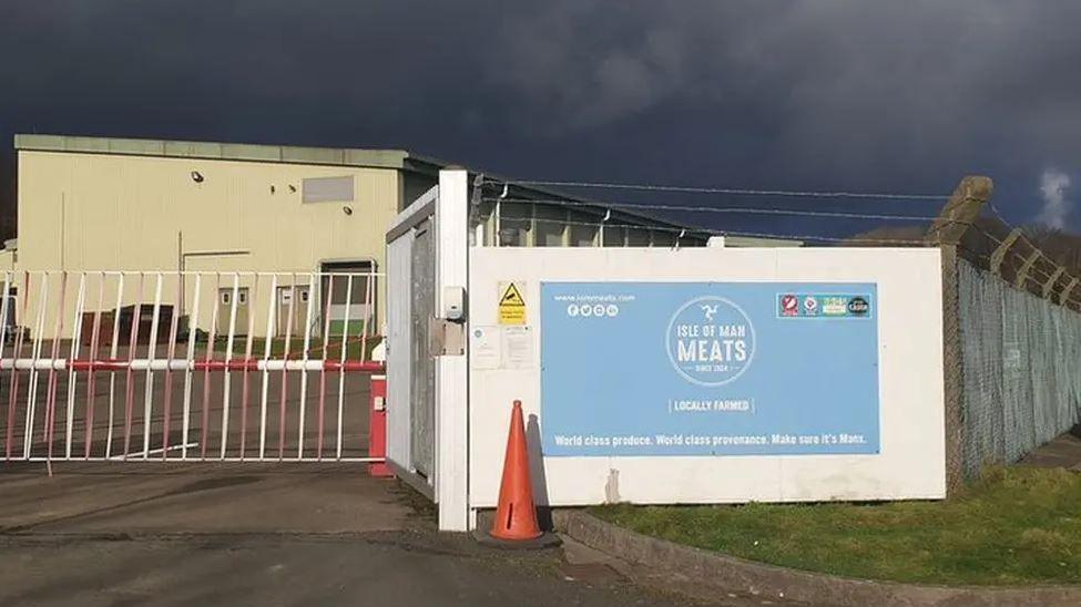 The meat plant, which is cream-coloured building with a flat roof. It has fencing topped with barbed wire around it and a red and white metal fence an a pale blue sign with the Isle of Man Meats logo in front of it.