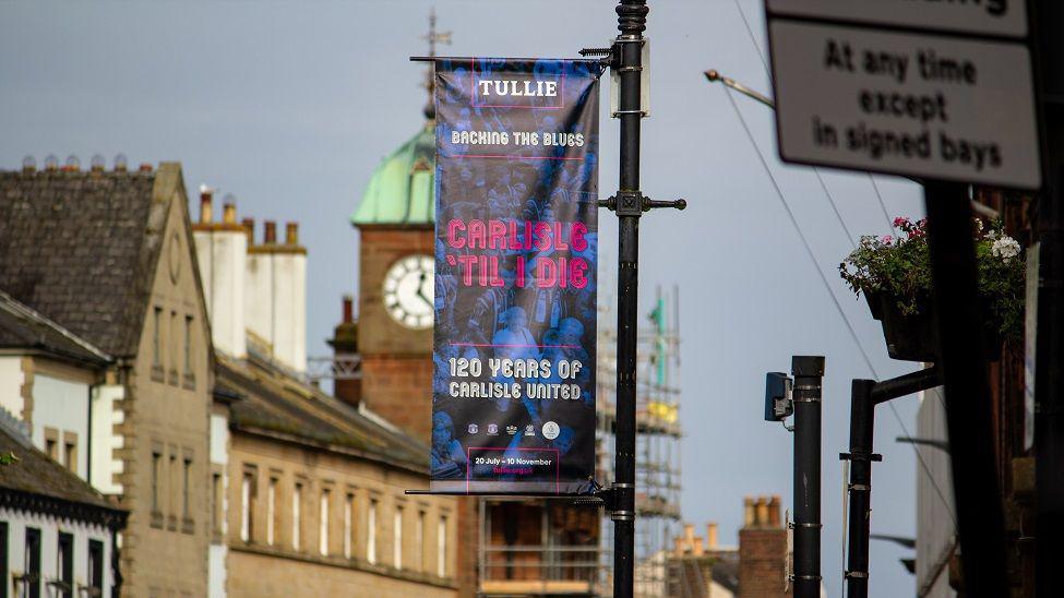 A blue and pink banner which reads 'Tullie- Backing the Blues- Carlisle 'Til I Die- 120 Years of Carlisle United'.