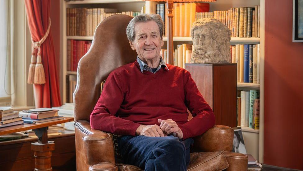 A man with grey hair sitting in a brown leather chair with his hands in his lap. He is wearing a red jumper and sat in front of a bookshelf and stone bust. 