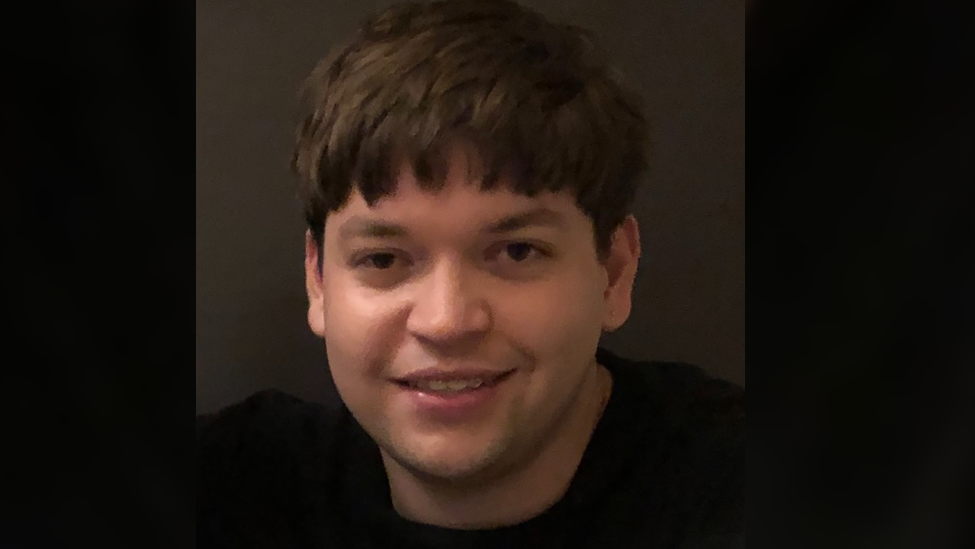 Luis Piovesana smiling at the camera. He has short brown hair with a fringe that reaches just above his eyebrows. He is wearing a black top.