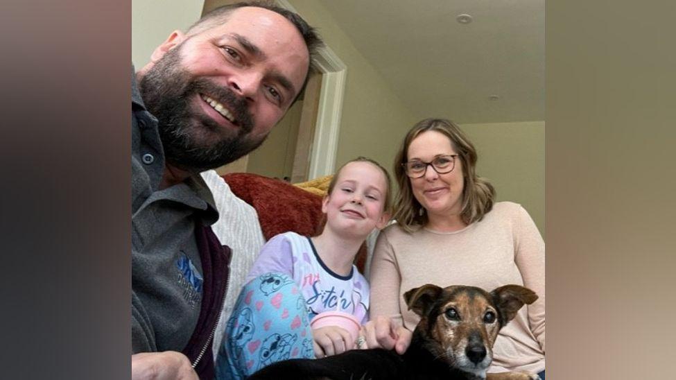 A man with a dark brown beard sits on a sofa next to a young girl in a pink top and a woman in a pale pink top. There is also a black and brown dog on the sofa.