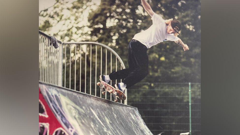 Ralph is skateboarding on a ramp. He is wearing a white shirt and black trousers and a cap back to front