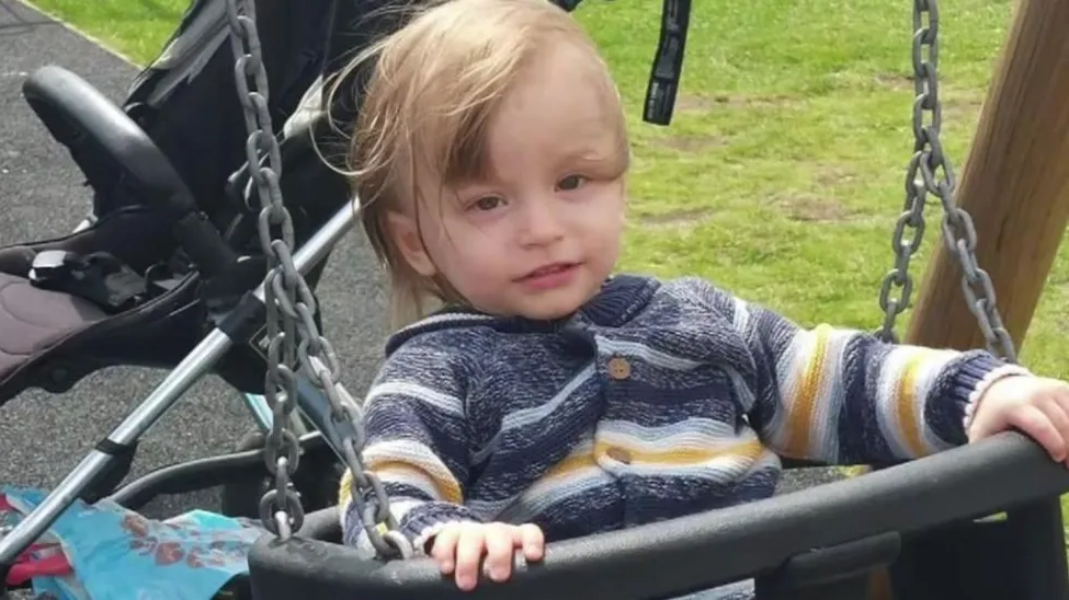 Bronson Battersby sits in a swing at the park. He is wearing a blue, white and yellow striped jumper. A pushchair is behind him.